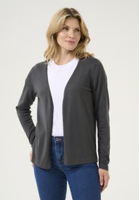 Šedý cardigan z měkké a lehké látky; otevřený přední design s dlouhými rukávy a volným střihem, skombinovaný s bílým tričkem a modrými džíny.