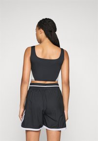 Kvinne med flettehår iført en svart og hvit kort tanktopp og svarte treningsshorts med hvite detaljer, vendt bort fra kameraet mot en enkel bakgrunn.