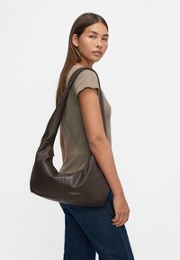 Giovane donna con lunghi capelli castani, che indossa una maglietta corta color taupe e jeans scuri, mentre porta una grande borsa a tracolla in pelle marrone scuro.