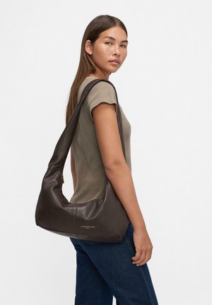 Giovane donna con lunghi capelli castani, che indossa una maglietta corta color taupe e jeans scuri, mentre porta una grande borsa a tracolla in pelle marrone scuro.