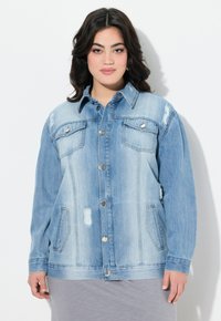 Vaaleansininen farkkutakki, jossa on napitettava etuosa, kaksi rintataskua ja kuluneita yksityiskohtia. Sisältää levitetyn kauluksen ja käännetyt hihat.