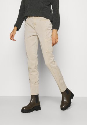 Henkilö, joka pukeutuu beigeihin cargohousuihin, tummanharmaaseen neuleeseen ja tummanruskeisiin, paksupohjaisiin nilkkureihin, seisoo yksinkertaista taustaa vasten.