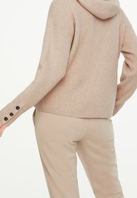 Hoodie en tricot côtelé beige avec une coupe légèrement raccourcie, doté d'une capuche proéminente et de boutons aux poignets. Associé à un pantalon beige.