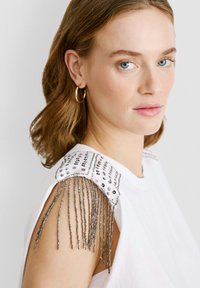 Valge õlatu top, millel on helmesõlmega disain ja hõbedased frantsiidetailid, luues tekstuuriga ja kihilise välimuse.