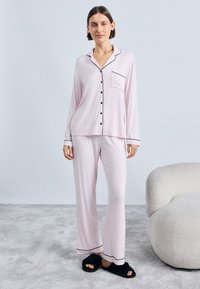 Set de pijama roz deschis cu piping negru; cămașă cu mâneci lungi și nasturi, cu buzunar și pantaloni asortați. Material moale și croială relaxată. Papuci negri.