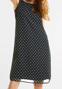 Svart ärmlös klänning med ett prickigt mönster i vitt. Tyget verkar lätt och mjukt, med en avslappnad passform och en knälång design.