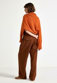 Kvinna med kort hår som bär en orange tröja över axlarna, bruna vida byxor och beige skor, stående mot en enkel bakgrund.