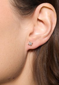 Złote kwadratowe kolczyki wkrętki z centralnym kamieniem z cyrkonią, otoczone małymi złotymi akcentami, na neutralnym kolorze skóry.