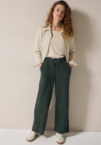 Světlý béžový sak, krémový top a tmavě zelené široké kalhoty se strukturovaným povrchem a stahovací šňůrkou v pase. Bílé tenisky dotvářejí outfit.