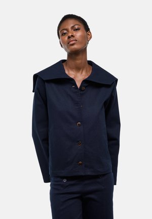 Femme portant une veste bleu marine foncé avec un grand col et des boutons, assortie à un pantalon bleu marine, debout devant un fond blanc.