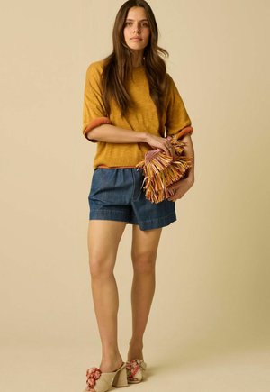 Vrouw met mosterdgele top en spijkershorts die een meerkleurige fringed clutch vasthoudt tegen een beige achtergrond.