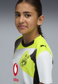 Camiseta de fútbol del Borussia Dortmund en blanco, negro y amarillo vibrante con el logo del patrocinador. Presenta mangas cortas, escudo del equipo y logo de Puma.