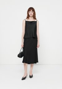 Svart satin camisole topp med tynne stropper, parret med et matchende svart knelengde skjørt. Modellen holder en liten svart håndveske og har på seg svarte mules.