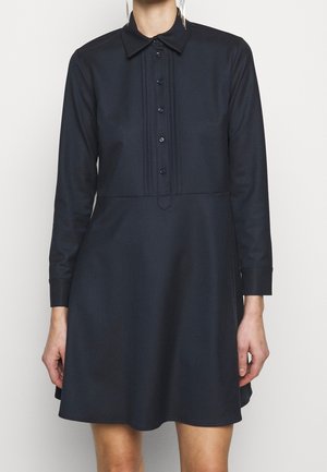 Femme portant une robe noire à manches longues avec col, boutonnière à l'avant et jupe évasée, se tenant devant un fond clair.