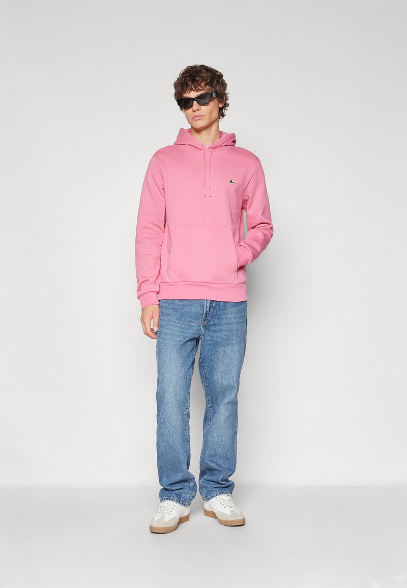 Sweat-shirt rose avec une poche avant et un cordon de serrage, associé à un jean bleu et des baskets blanches, sur un fond uni.