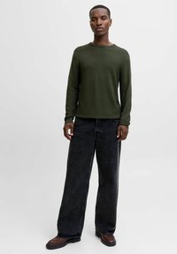 Cămășă verde măsliniu cu mânecă lungă, cu un model rib și purtată cu pantaloni din denim negri, lejeri, și încălțăminte marron. Design simplu, cu țesătură texturată.