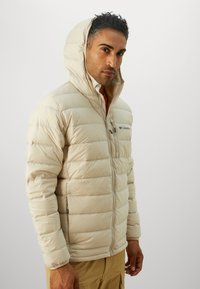 Beige puffer jacket with a hood, featuring quilted design, front zipper, and a left chest pocket. Material appears smooth and insulated.