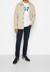 Beige bomulljacka över en vit T-shirt med ett blått fjärils-motiv, matchat med mörk denimjeans och svarta sneakers med vita detaljer.