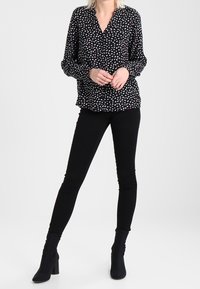 Blusa nera a maniche lunghe con un motivo astratto bianco e rosso, scollatura a V e vestibilità comoda, abbinata a pantaloni skinny neri e stivaletti.