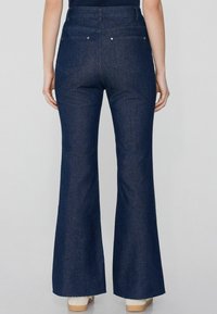 Jeans acampanados de un azul oscuro con cintura alta, que cuentan con cinco bolsillos, herrajes plateados y un dobladillo deshilachado en la parte inferior, hechos de tela de mezclilla.
