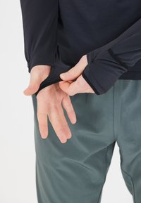 Langärmliges schwarzes Sportoberteil mit Daumenlöchern, aus glattem Material und mit figurbetontem Design, kombiniert mit grünen Outdoor-Hosen.