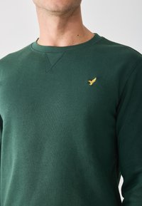 Zielony sweatshirt z okrągłym dekoltem, wykonany z miękkiego materiału. Posiada małe, złote logo ptaka na klatce piersiowej oraz prosty, prążkowany dekolt.
