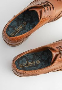 Zapatos de cuero marrón con detalles de brogue, que cuentan con una plantilla de patrón floral oscuro y cordones negros, exhibidos desde arriba.