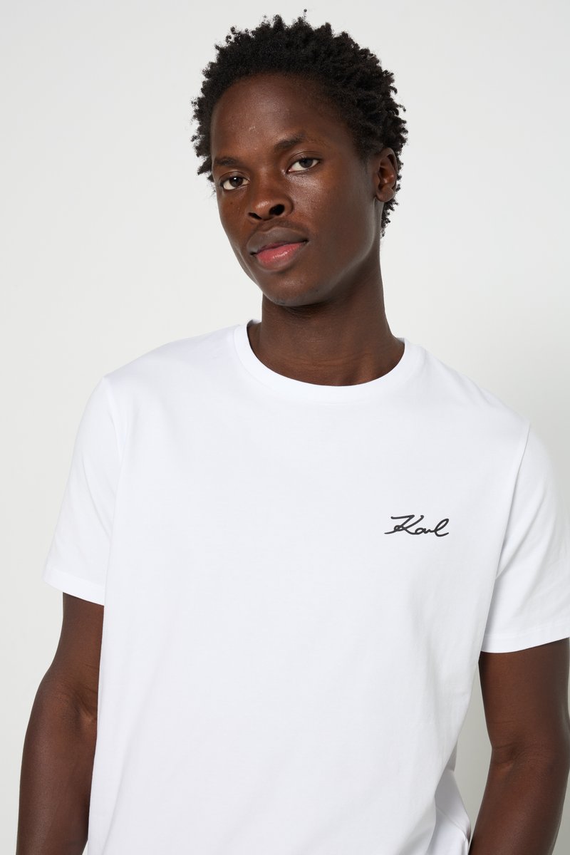 Camiseta de algodón blanca con cuello redondo, mangas cortas y un logo en script negro en la parte izquierda del pecho. Textura suave, corte relajado.