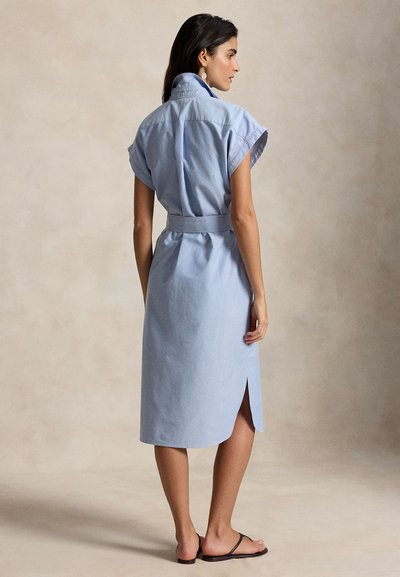 Γυναίκα με ανοιχτό μπλε μεσοφόρεμα με ζώνη, μανίκια καπέλο και ανοίγματα στην πλάτη, στέκεται ξυπόλυτη σε μπεζ πάτωμα με ουδέτερο φόντο.