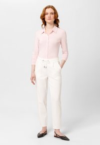 Camicia a maniche lunghe rosa chiaro con colletto, abbinata a pantaloni bianchi con cordoncino. Il modello indossa scarpe basse scure. Design semplice e aderente.