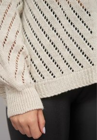 Beige gestrickter Pullover mit einem Lochmuster und diagonalen Streifen, gerippten Bündchen und Saum. Texturiert, leichtes Material, eng anliegend an den Handgelenken.