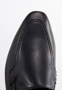 Chaussure en cuir noir avec un bout pointu et des coutures le long des côtés. Texture lisse, design formel et sans élément matériel visible.