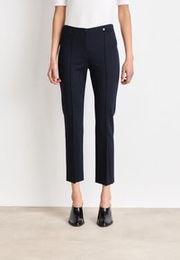 Pantaloni navy con una vestibilità sartoriale, dotati di chiusura a bottone e dettagli in piping laterale, che terminano poco sopra la caviglia. Abbinati a mule nere.