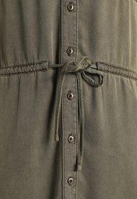 Gros plan sur un tissu vert olive avec des boutons à l'avant et un cordon noué à la taille.