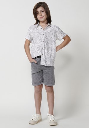 Camisa blanca de mangas cortas con un patrón de hojas grises, combinada con pantalones cortos de mezclilla grises. El calzado consiste en zapatillas beige con cordones blancos.