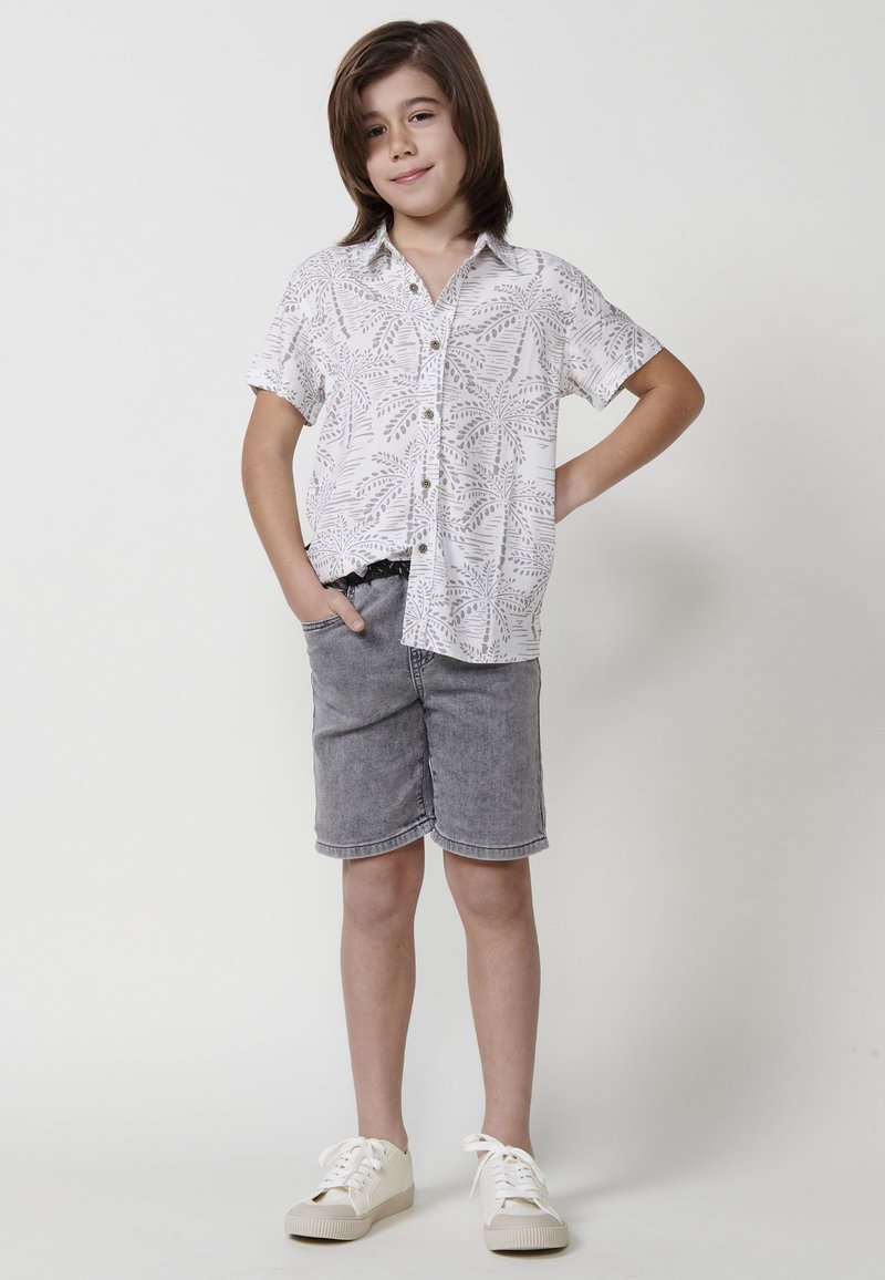 Kratkų rankovių balta marškinė su pilku lapų raštu, derinami su pilkais džinsiniais šortais. Avalynė – smėlio spalvos sportbačiai su baltais raišteliais.