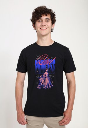 Giovane uomo che indossa una maglietta nera con testo "Rebel Princess" blu e rosso e grafica di una donna e un robot, pantaloni beige, sorridente, in piedi su sfondo bianco.