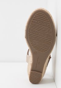 Semelle d'une sandale compensée beige avec semelle en caoutchouc texturée, marquage "Tom Tailor" et taille 37, inclinée contre une surface blanche.