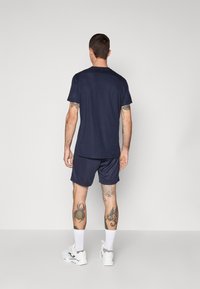 Marineblaue Sportbekleidung bestehend aus einem T-Shirt mit kurzen Ärmeln und Shorts, hergestellt aus einem glatten Stoff. Sneakers und sichtbare Tattoos an den Beinen.