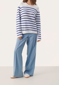 Maglione a righe bianco e blu, abbinato a pantaloni larghi azzurri. Il maglione ha maniche lunghe e collo rotondo; le scarpe sono bianche con suole in gomma.