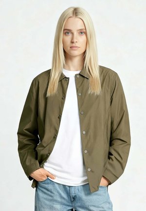 Vrouw met lang blond haar, gekleed in een olijfgroene jack met knopen over een wit shirt en lichtblauwe spijkerbroek, staand met handen in de zakken.