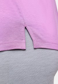 Lila, leichtes Hemd mit seitlichem Schlitzdetail, darüber getragen ein strukturiertes graues Kleidungsstück mit Rautenmuster.