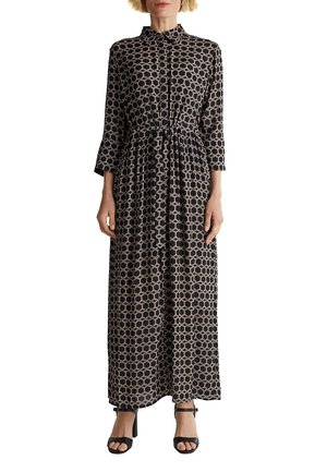 Kobieta w długiej sukni maxi z długim rękawem, czarno-beżowym wzorem, kołnierzem i wiązaniem w pasie, w zestawieniu z czarnymi sandałami na obcasie z odkrytymi palcami.