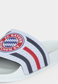 Άσπρη σαγιονάρα με το λογότυπο της FC Bayern München και κόκκινες και μαύρες ρίγες στο επάνω λουράκι.