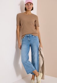 Femme portant un haut marron à manches courtes, un jean large bleu clair, des baskets beige et une casquette rose, tenant une veste beige.