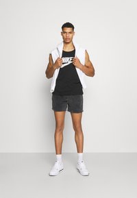 Sort Nike tank top, hvid ærmeløs hættetrøje, sorte denimshorts og hvide sneakers. Model står på en grå baggrund.