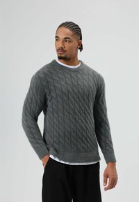 Grijze kabelgebreide trui met lange mouwen en een ronde hals, gemaakt van een zacht, gestructureerd materiaal, gedragen over een wit overhemd.