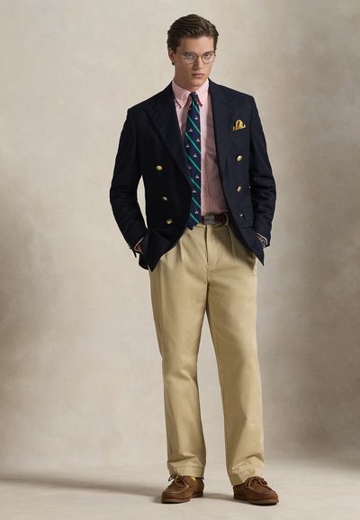 Jonge man met een ronde bril, marineblauw colbert met gouden knopen, roze overhemd, patroon stropdas, beige broek en bruine schoenen, staand met handen in de zakken.