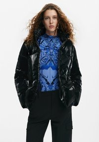 Giubbotto puffo nero con finitura lucida, cappuccio oversize e chiusura con zip, indossato sopra una maglietta blu a fantasia e pantaloni neri.