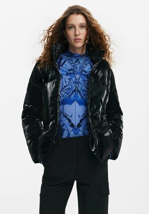 Giubbotto puffo nero con finitura lucida, cappuccio oversize e chiusura con zip, indossato sopra una maglietta blu a fantasia e pantaloni neri.
